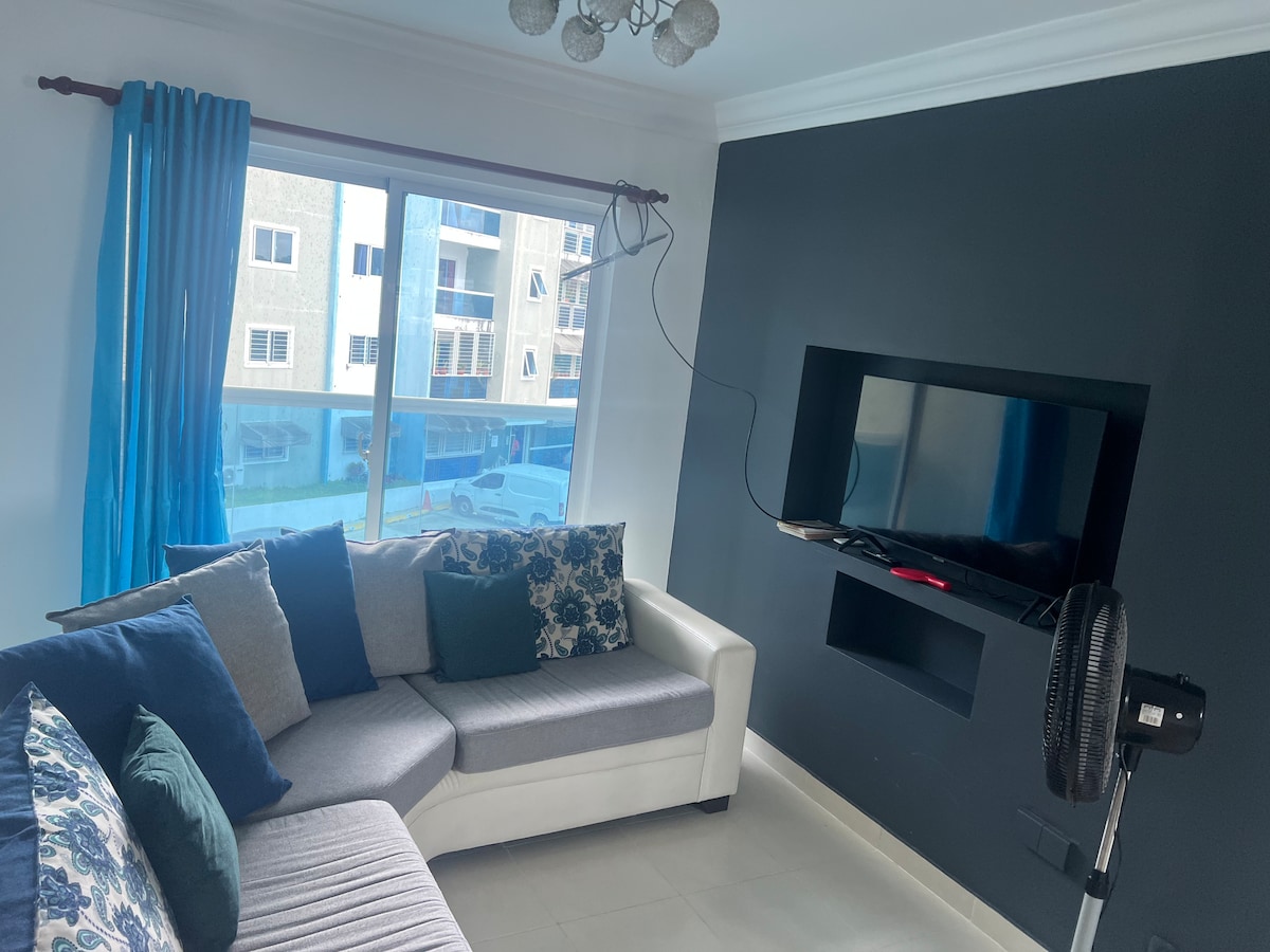 A modern living area features a sectional sofa adorned with decorative cushions. A flat-screen television is mounted on a dark wall, while large windows dressed in blue curtains offer views of the surrounding area. A standing fan is positioned nearby.