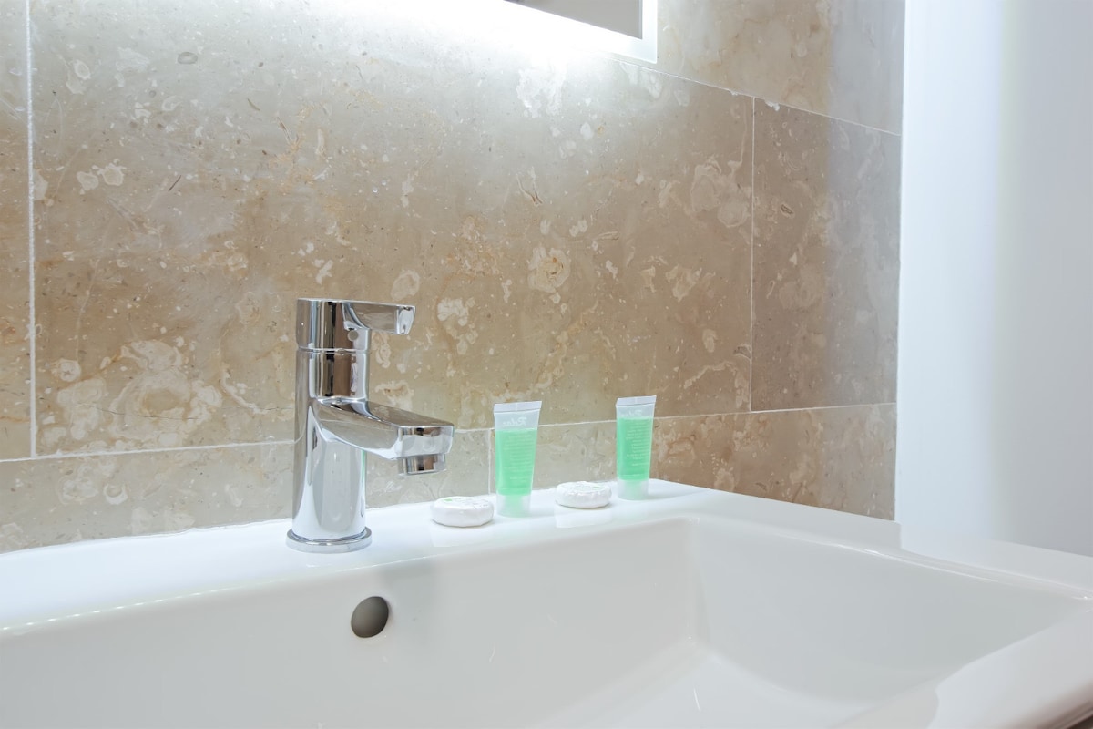 A clean bathroom sink is presented, featuring a modern chrome faucet. Two small green bottles and cotton pads are neatly arranged on the countertop. The wall is adorned with textured beige tiles, complementing the bright, minimalist design of the space.