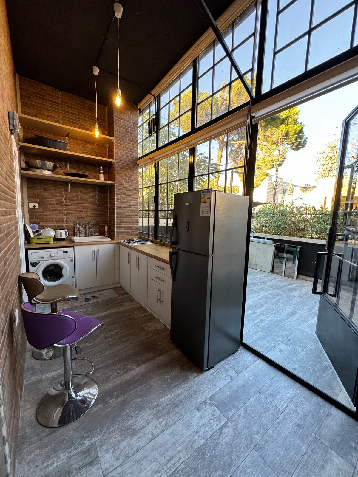 A modern kitchen area features sleek cabinetry and open shelving, complemented by large windows that provide ample natural light. A stainless steel refrigerator and washing machine are present, alongside a high-backed bar stool in purple. The door opens to a balcony with greenery.