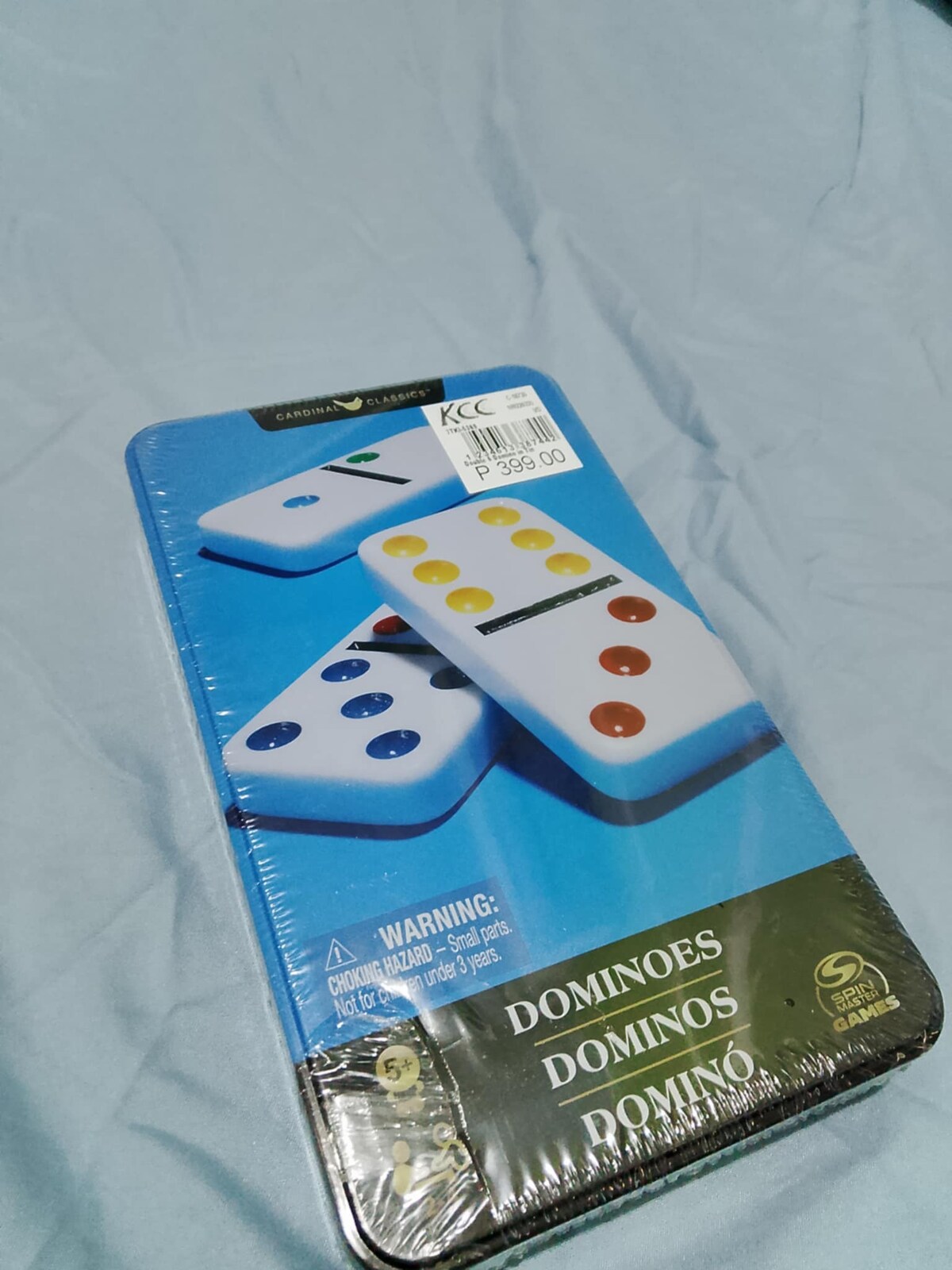A sealed box of dominoes is displayed on a light blue fabric surface. The box features images of colorful domino pieces in blue, yellow, and white. The label on the box indicates that it is a game, with a warning for small parts.