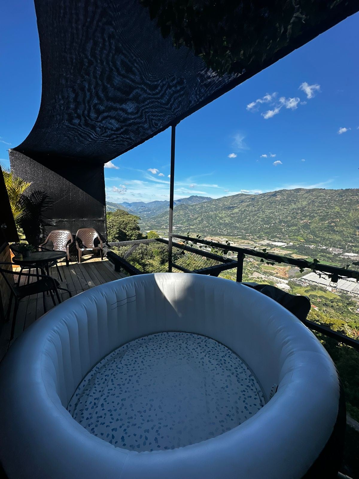 A private outdoor space features a circular inflatable hot tub surrounded by wooden decking. Comfortable chairs are positioned nearby, and a shaded area is created by an awning. Stunning mountain views and blue skies are visible in the background.