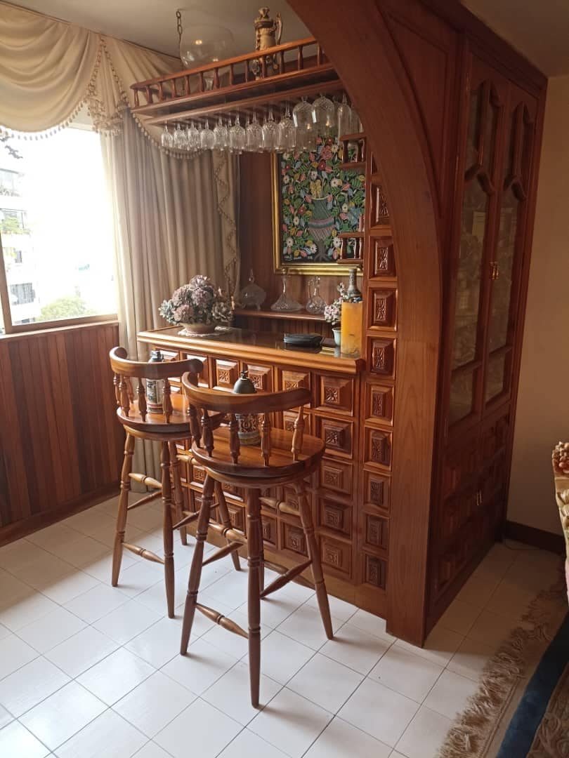 A wooden bar area is featured, showcasing intricately carved panels and a glass-front cabinet. Two high wooden stools are positioned at the bar. Natural light enters through a nearby window, highlighting the warm tones of the wood and the decorative details.