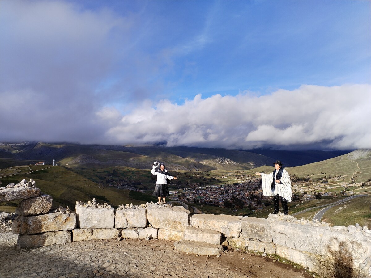 A scenic view encompasses rolling hills under a partly cloudy sky. Two individuals are positioned on a stone wall, one wearing a traditional outfit and the other holding a blanket. A small town is visible in the valley below, framed by mountainous terrain.