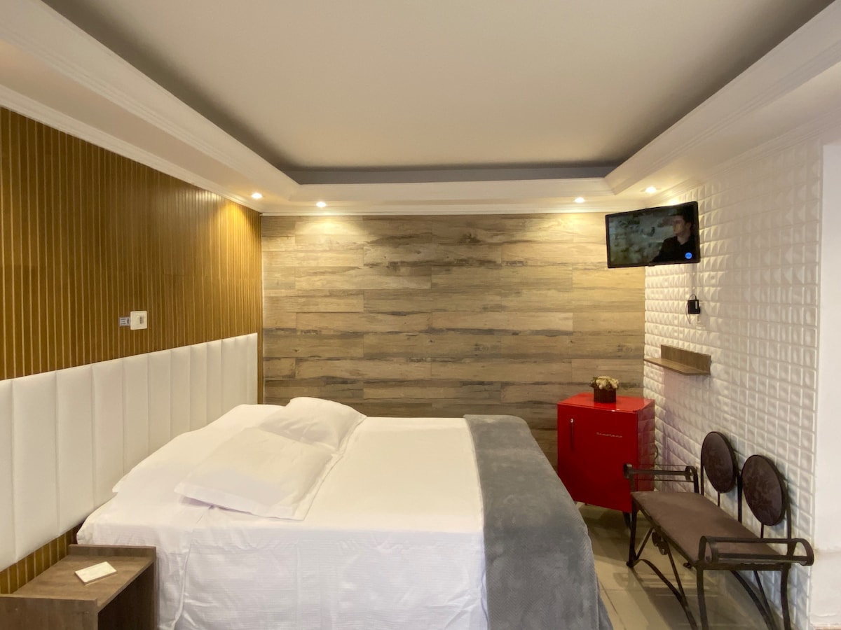 A contemporary bedroom is featured, showcasing a spacious bed with white linens and a light gray throw. Accents of wood and textured walls create a modern aesthetic. A red cabinet and seating area provide functionality, while a wall-mounted TV enhances entertainment options.