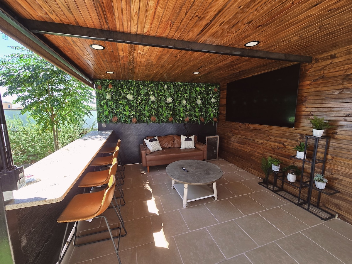 An outdoor seating area features a wooden ceiling and wall adorned with greenery. A sleek bar counter with high stools is complemented by a cozy leather sofa. A modern coffee table sits in front, while a flat-screen television is mounted on a wooden panel.