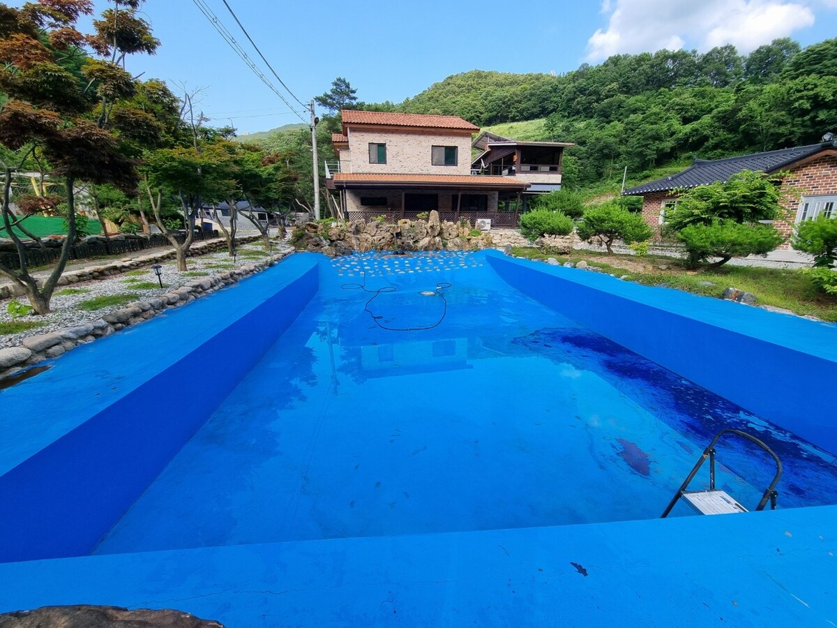 A vibrant blue swimming pool is surrounded by stone pathways and lush greenery. The pool area is bordered by neatly trimmed bushes, providing a sense of privacy. In the background, a two-story building is visible, set against a hillside of trees.