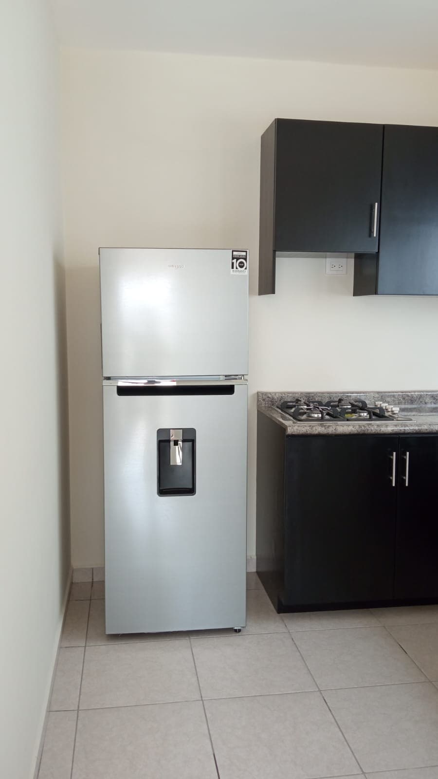 A modern kitchen space contains a sleek silver refrigerator with an ice and water dispenser. Dark cabinetry complements the granite countertop, alongside a gas stove with multiple burners. Neutral-colored tiles extend across the floor, enhancing the kitchen's clean and functional look.