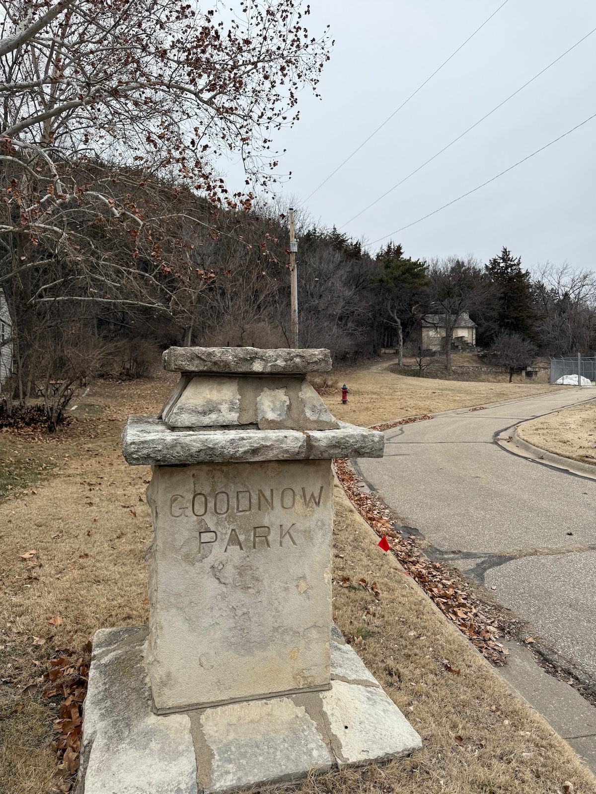 A weathered stone monument displays the name 'Goodnow Park,' standing beside a winding road. Surrounding trees are bare, indicative of the season. The landscape features a gentle hill in the background, partially obscured by foliage, and a gravel pathway leading towards the park.