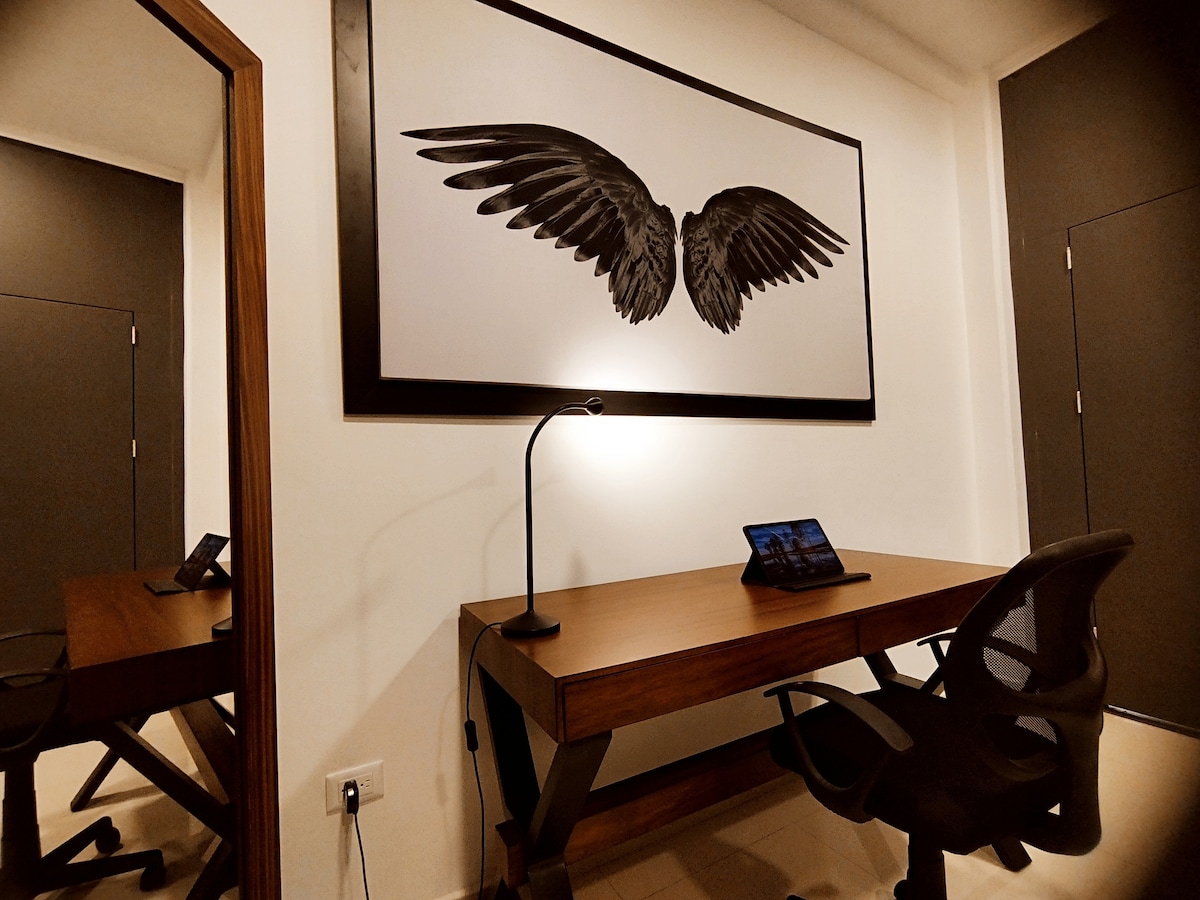 A workspace is shown, featuring a wooden desk and an ergonomic chair. A modern lamp provides focused lighting, while a tablet rests on the desk. A large artwork of stylized wings is mounted on the wall, adding visual interest to the room.