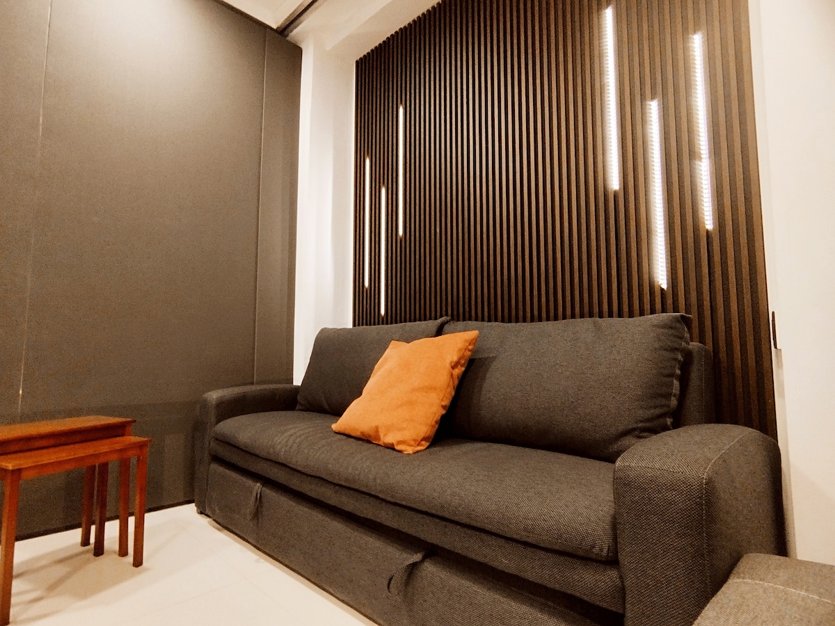 A modern lounge area features a spacious gray sofa adorned with an orange accent pillow. A small wooden table is positioned in front of the sofa. A textured wooden wall provides a unique backdrop with vertical lighting fixtures creating subtle highlights.