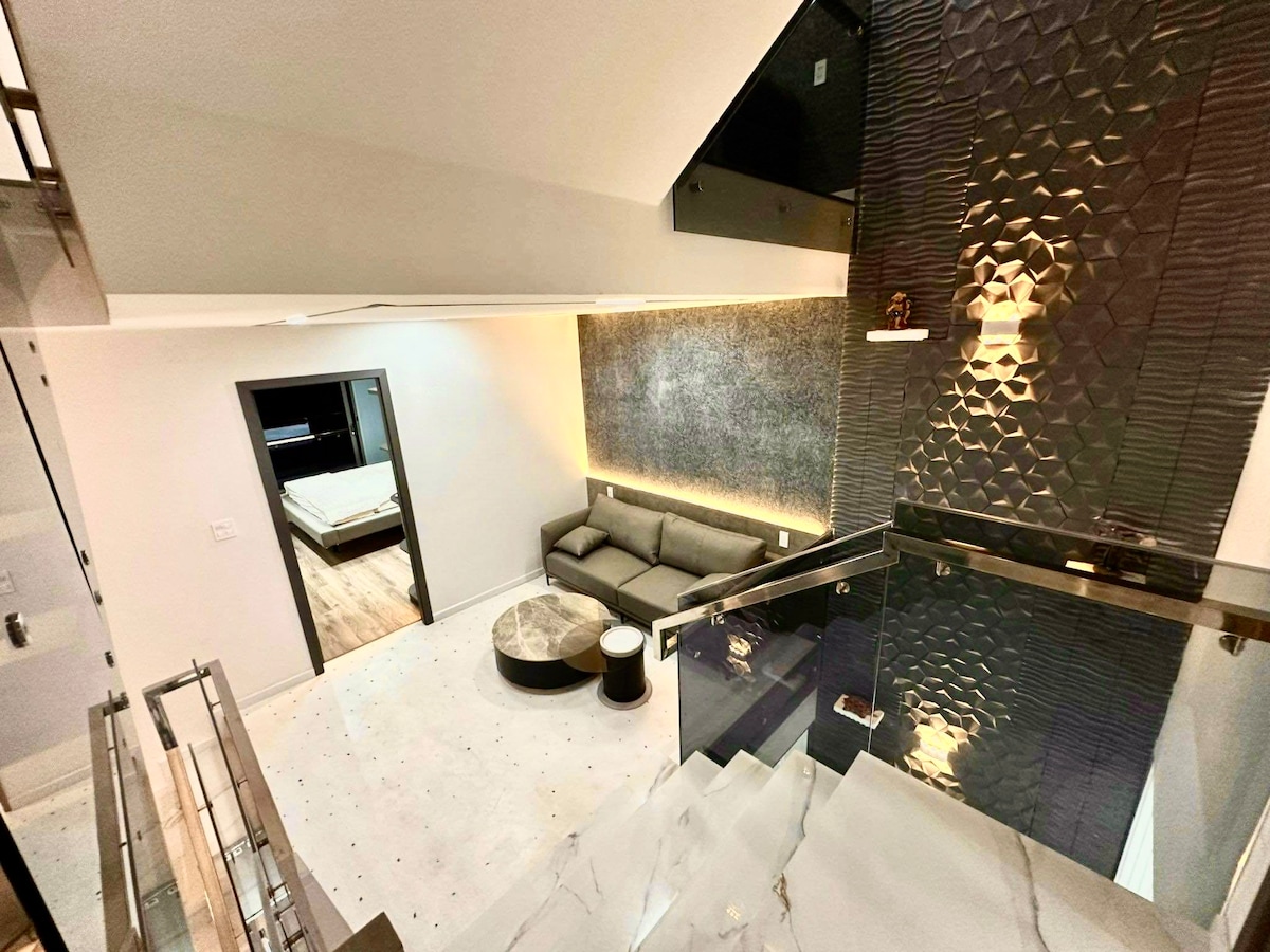 An open living area features a modern sofa and a round coffee table, with soft lighting accentuating the textured walls. A staircase with glass railings leads to the upper level, where a glimpse of a spacious bedroom can be seen through a doorway.