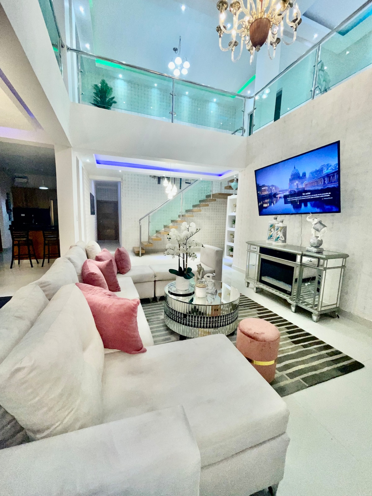 A spacious living area is presented with a light-colored sectional sofa adorned with pink cushions. A glass coffee table sits atop a striped rug, while a decorative piece is displayed on a contemporary console table. Bright lighting enhances the open layout, complemented by a large television mounted on the wall.