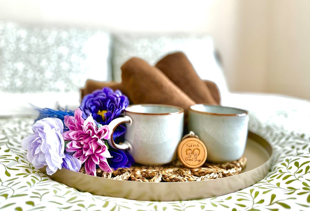 A tray holds two ceramic cups alongside a wooden charm and an arrangement of colorful artificial flowers. Soft brown towels are placed neatly in the background on the bed, which features a patterned duvet and pillow for a cozy touch.