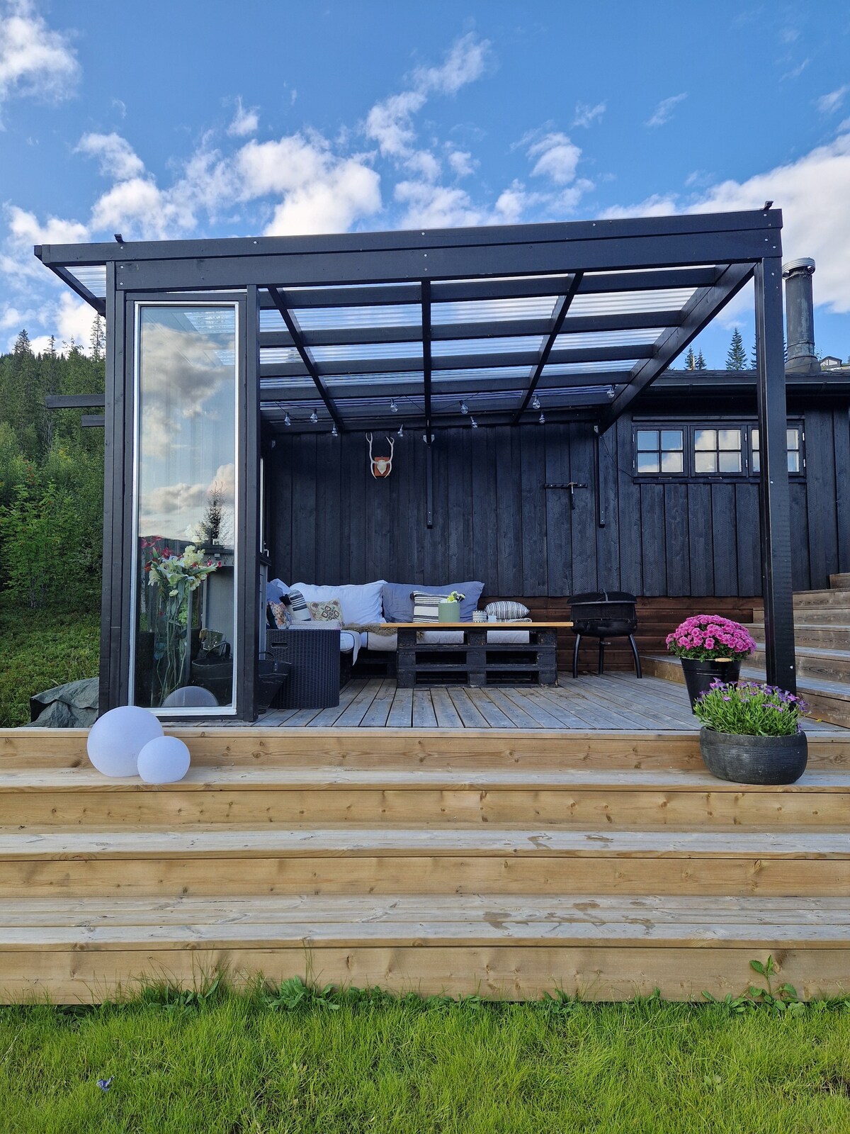 A spacious terrace with a canopy provides a comfortable seating area, featuring a sectional sofa and a coffee table. Potted flowers add a touch of color. Steps lead down to a green lawn, framed by trees and the natural landscape.