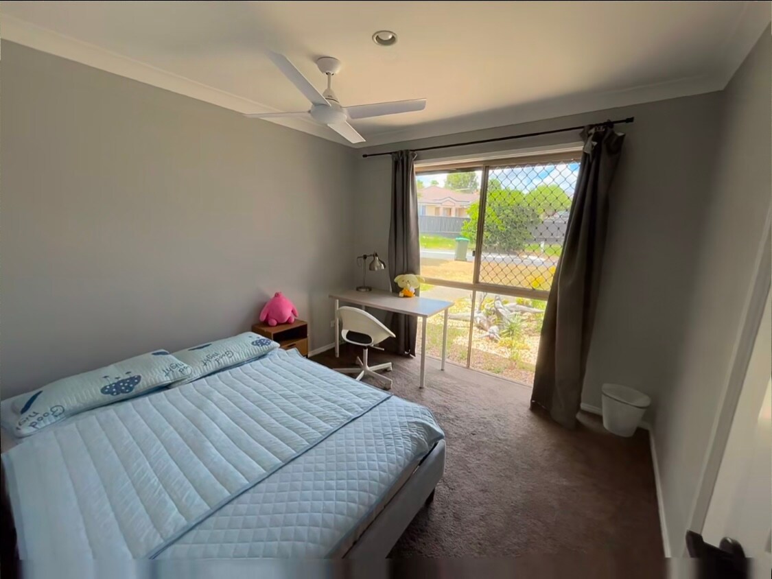 A quiet bedroom features a spacious bed with a light-colored quilt. A window provides natural light and a view of the outdoors, complemented by dark curtains. A small desk and chair are positioned nearby, adding functionality to the room.