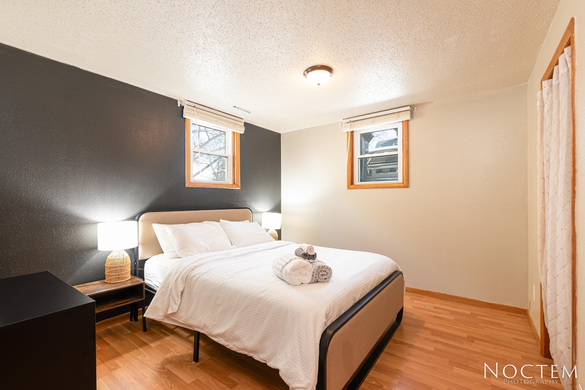 A cozy bedroom is furnished with a spacious bed dressed in white linens. Soft lighting is provided by two bedside lamps on woven bases. A nightstand sits beside the bed, and two windows are covered with light window treatments, allowing natural light to filter in.