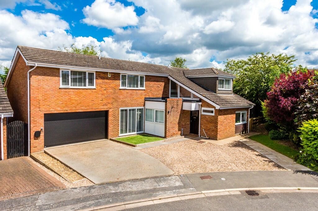 The exterior of a spacious two-story home is depicted, featuring a combination of brick and a sleek facade. A long driveway extends to the front, accommodating several vehicles. Lush greenery surrounds the property, with trees and shrubs providing a natural border.