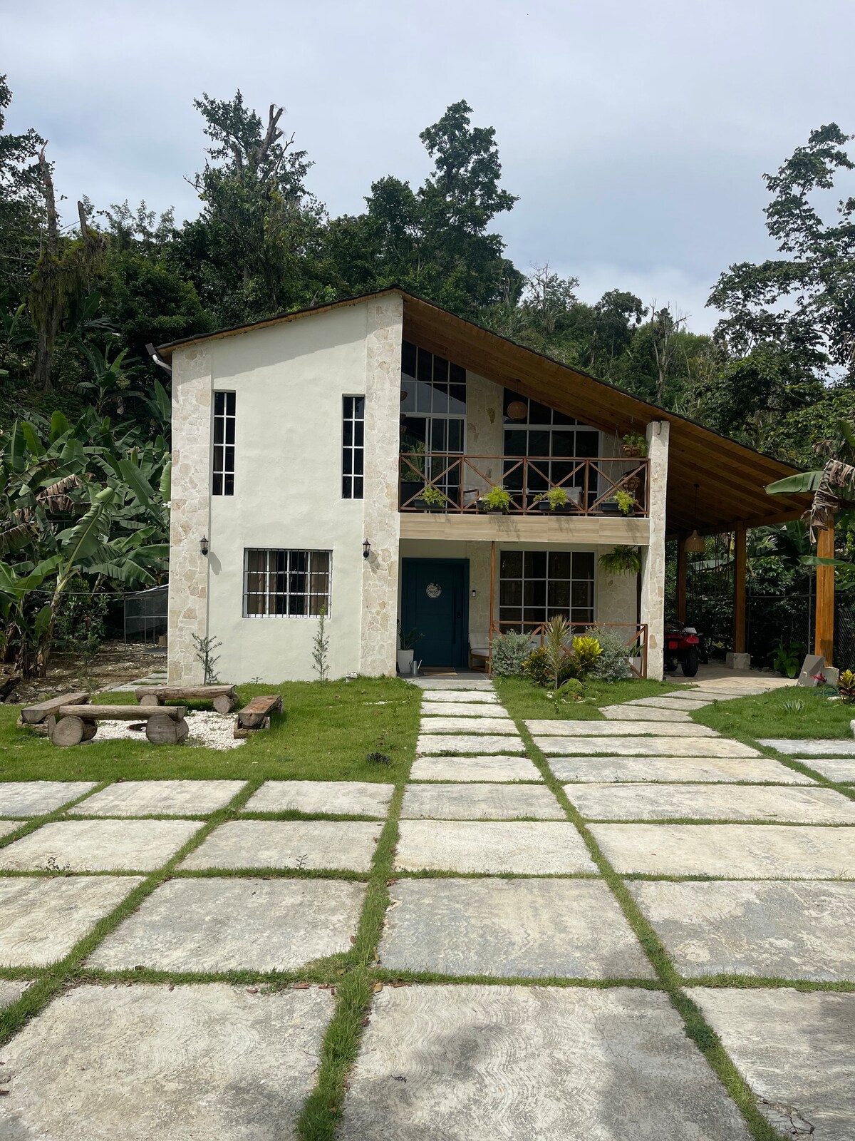 A two-story house with a light-colored exterior features large windows, allowing natural light to enter. A covered porch extends from the front, and a stone path leads to the entrance. Lush greenery surrounds the property, creating a serene outdoor space.