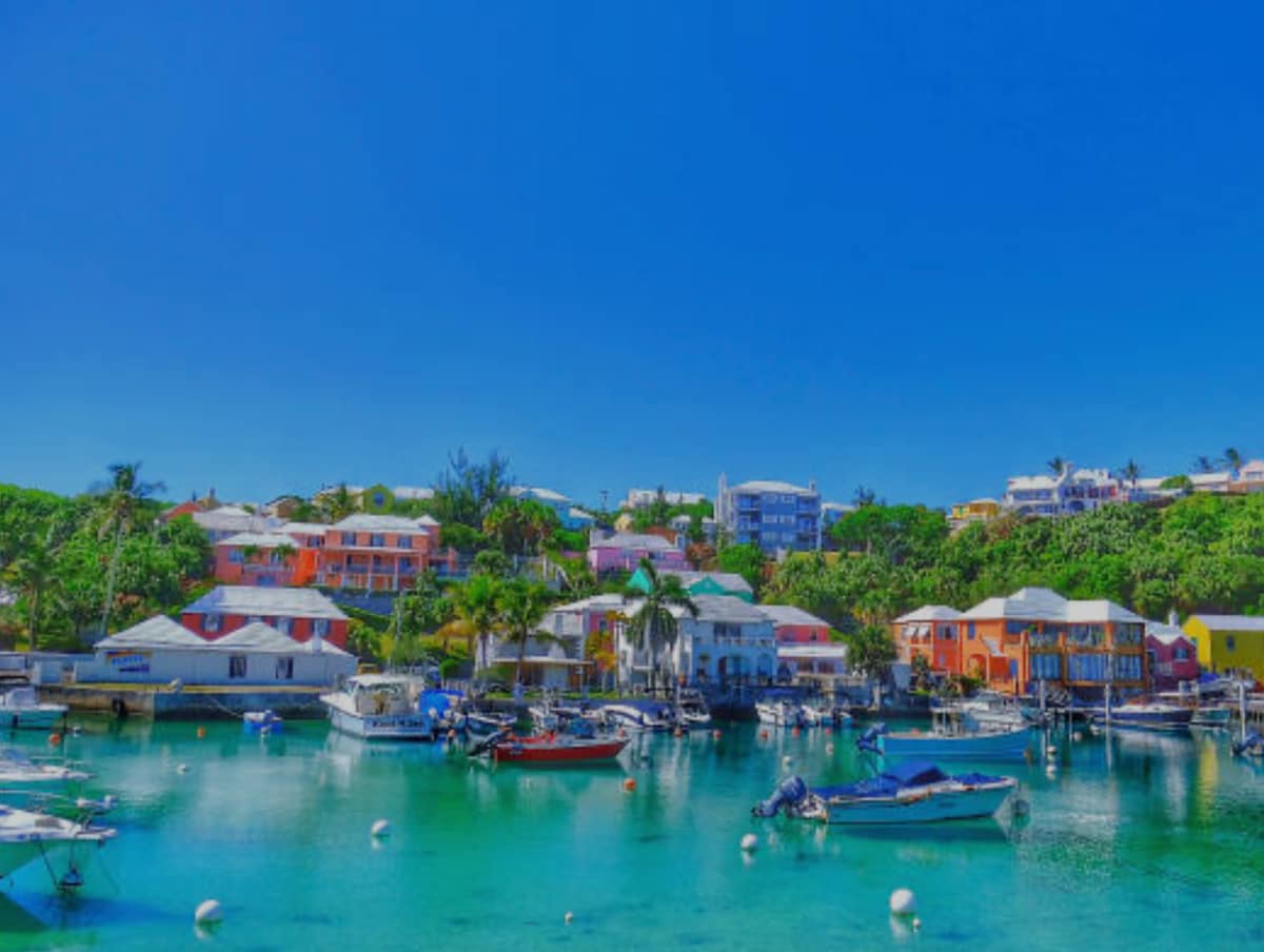 A vibrant waterfront scene features colorful buildings nestled along a serene harbor, with boats gently bobbing in the turquoise water. Lush greenery surrounds the area, and the clear blue sky enhances the inviting atmosphere of this picturesque harborfront location.