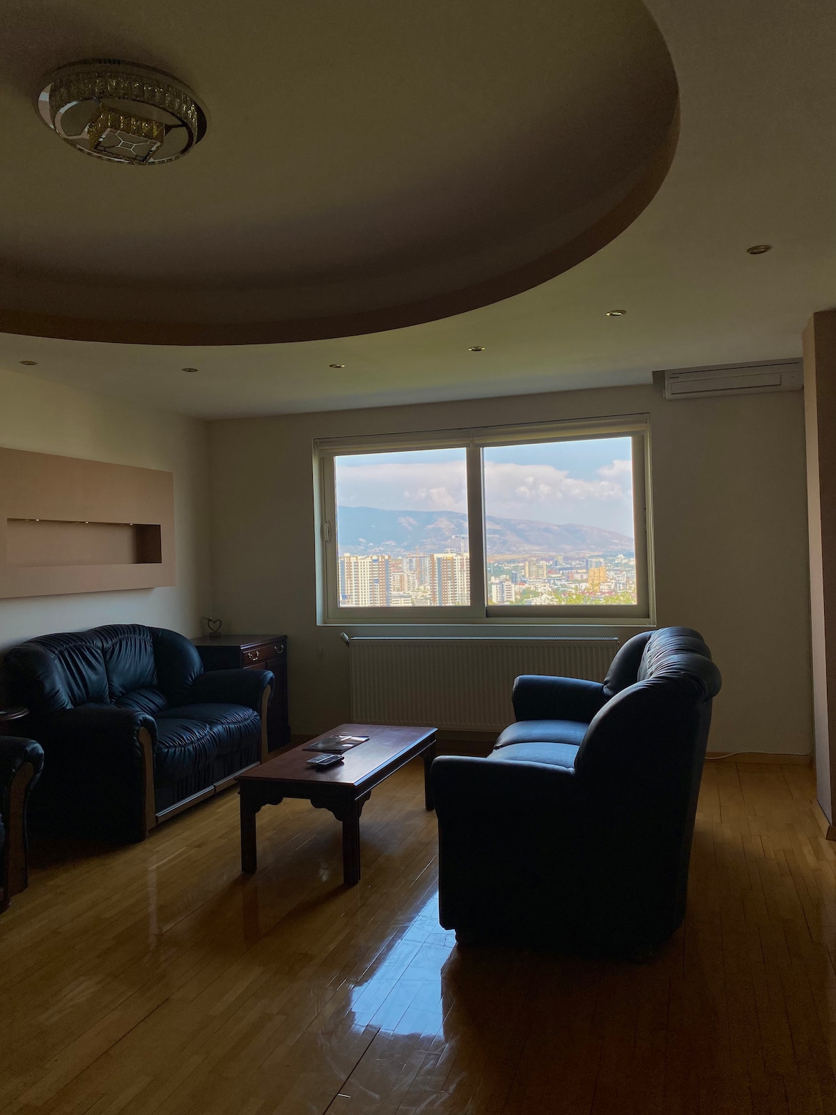 A cozy living area features two black leather chairs facing a wooden coffee table. A large window offers panoramic views of the city and surrounding mountains, allowing natural light to illuminate the space. The ceiling is adorned with a decorative fixture, enhancing the room's ambiance.
