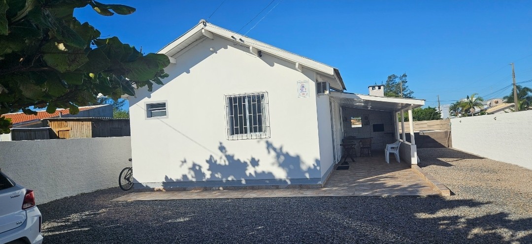 A single-story house with a white exterior is surrounded by a gravel driveway. A shaded entrance is visible, featuring simple outdoor furniture. A bicycle is parked nearby, contributing to a relaxed outdoor environment with clear blue skies overhead.
