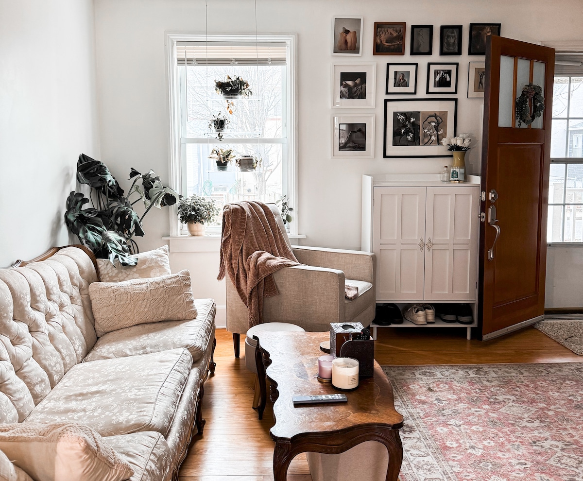 The living room features a comfortable sofa and an armchair paired with a wooden coffee table. Plants hang in the window, adding natural elements. A collection of framed photos decorates the wall, while a door with a glass panel provides exterior access.