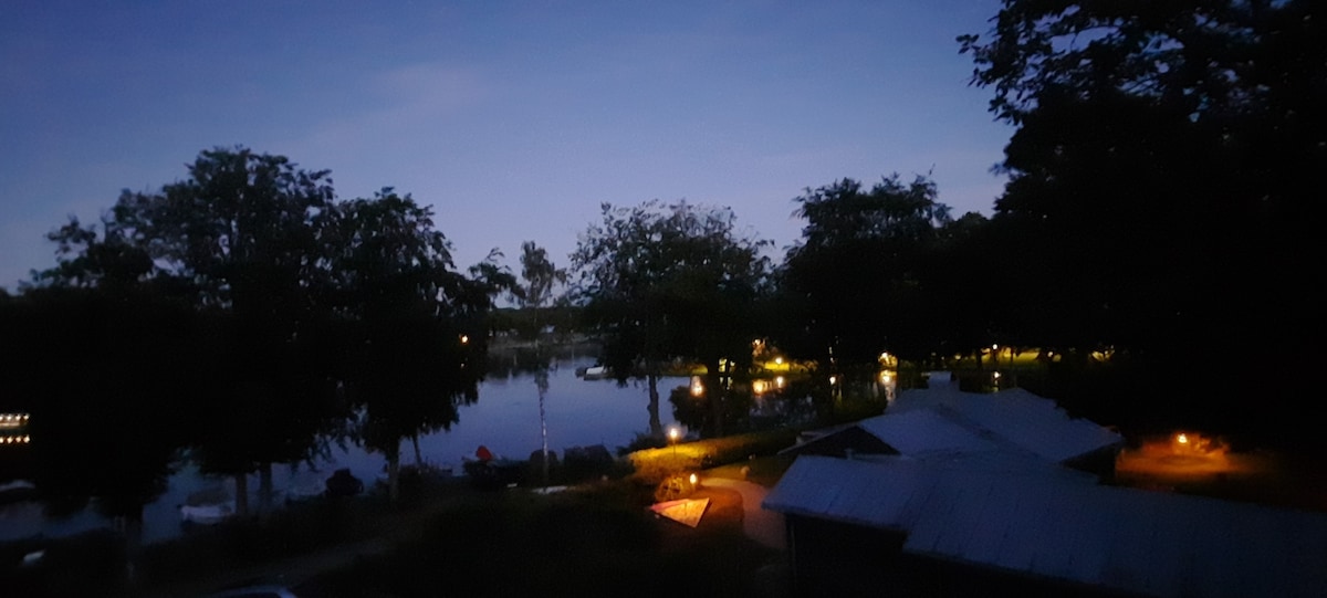 A serene waterfront view is captured at dusk, showcasing calm waters reflecting soft illuminations from nearby trees and structures. Silhouetted outlines of foliage and buildings create a tranquil atmosphere, while the gentle glow of lights enhances the peaceful evening setting.