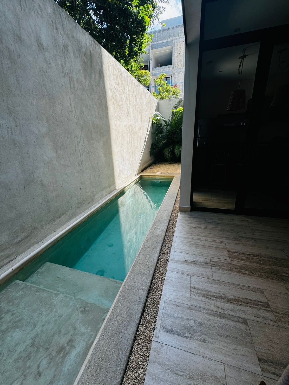 A narrow private pool is visible, reflecting the sunlight on its serene surface. Surrounding greenery adds a natural touch, while smooth stone and textured tile line the pool area. Glass doors are positioned nearby, connecting the outdoor space to the interior.