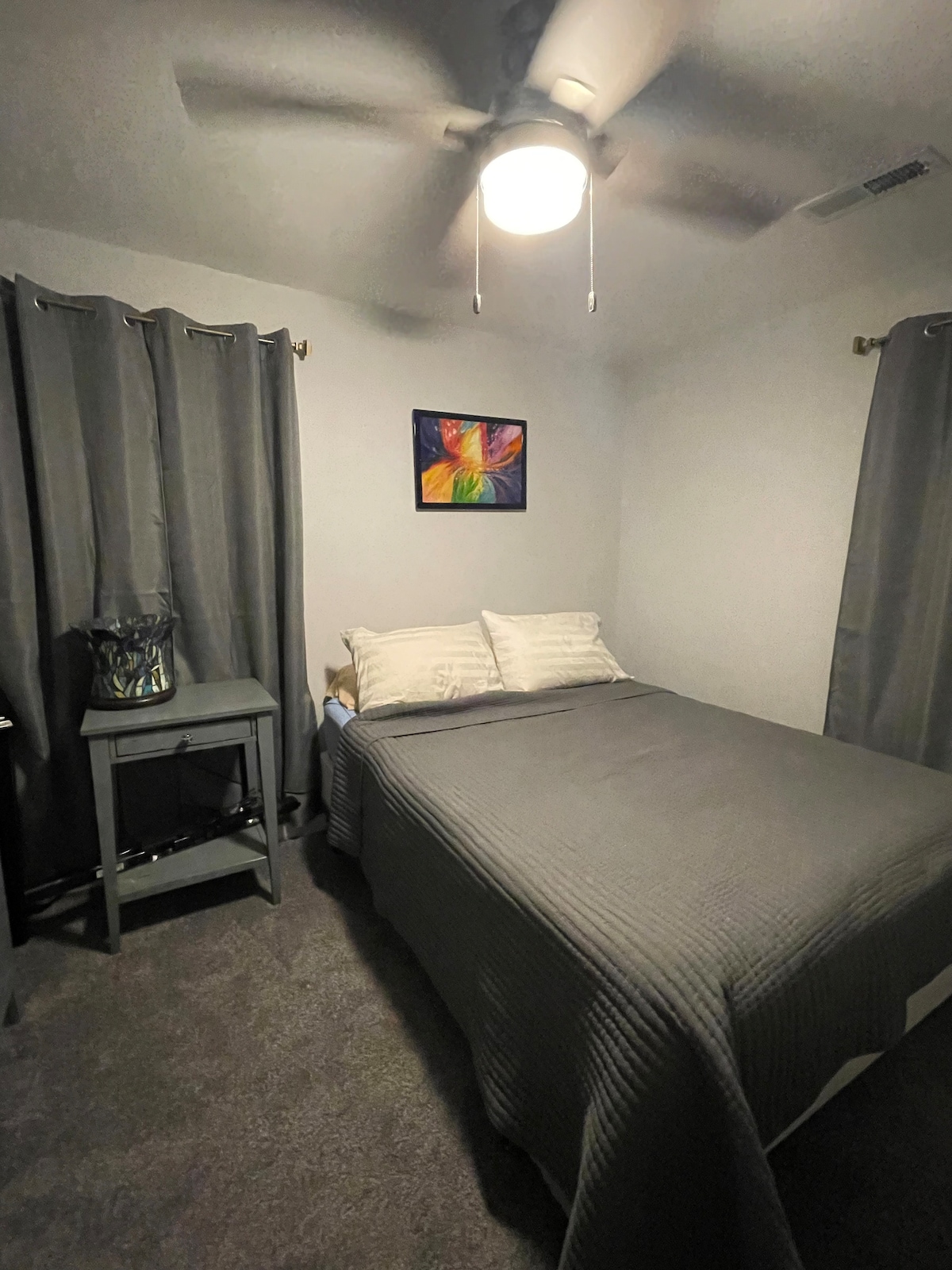 A cozy bedroom features a spacious bed with light gray bedding and two pillows. A painting with vibrant colors is displayed on the wall above. Curtains frame a window, and a bedside table holds a small decorative basket.