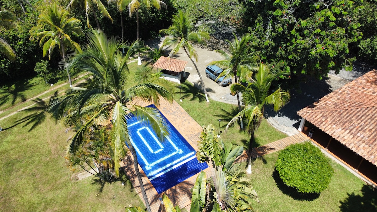 An aerial view reveals a bright blue swimming pool surrounded by lush green grass and palm trees. A rustic structure with a tiled roof is visible nearby, along with a gravel pathway leading to a parked vehicle.