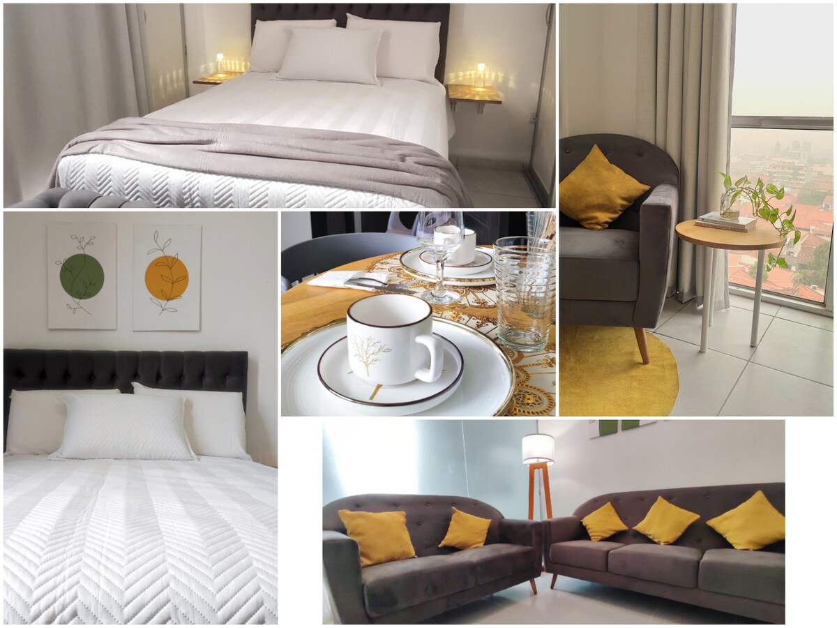 The image features a cozy bedroom with a spacious bed dressed in crisp white linens and light grey accents. A comfortable seating area is visible, showcasing plush chairs with decorative yellow cushions. A dining table is set with elegant tableware, complemented by decorative wall art.