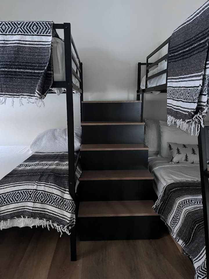 غرفة ذات سريرين علويين وسريرين مفردين مع مكتب/منطقة مكتب صغيرة وسلالم للدخول والخروج من السرير العلوي بسهولة.