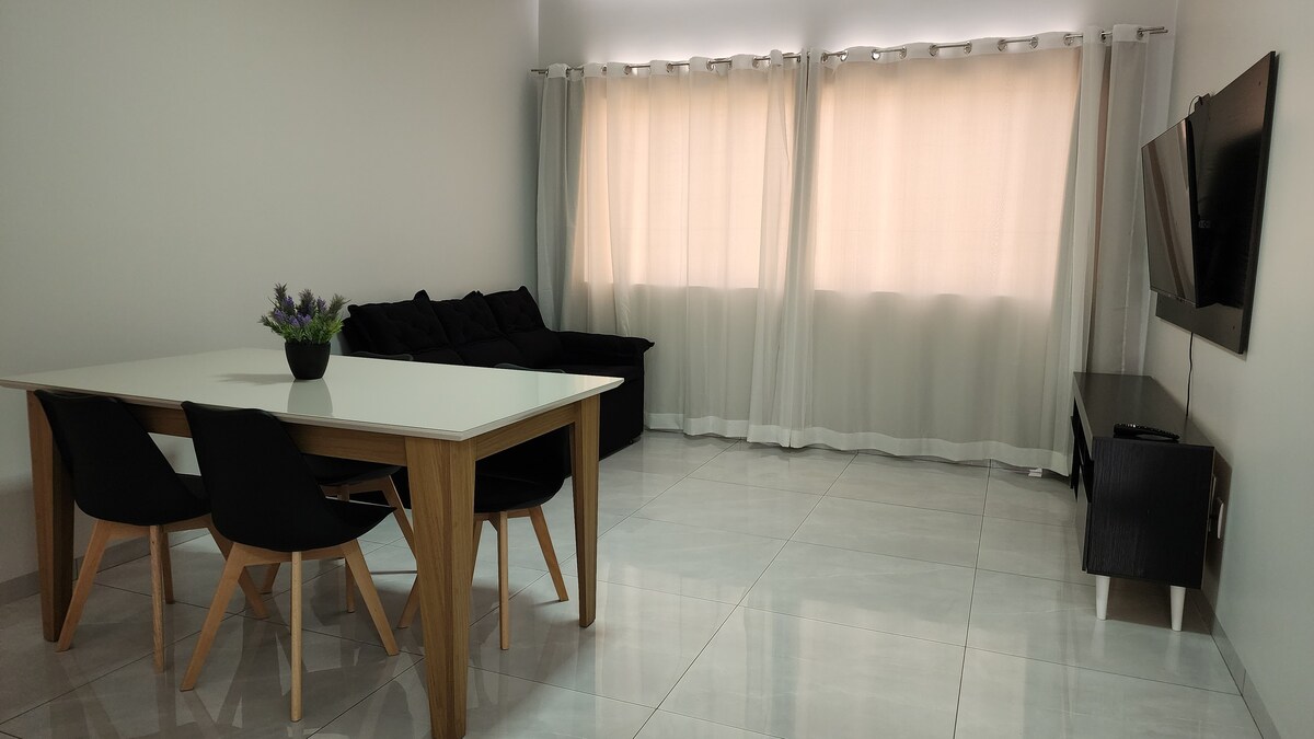 A bright living area features a white dining table with four black chairs. A dark sofa is positioned against the wall, while a mounted television is present across from the seating area. Light filters through sheer curtains, adding a soft glow to the space.