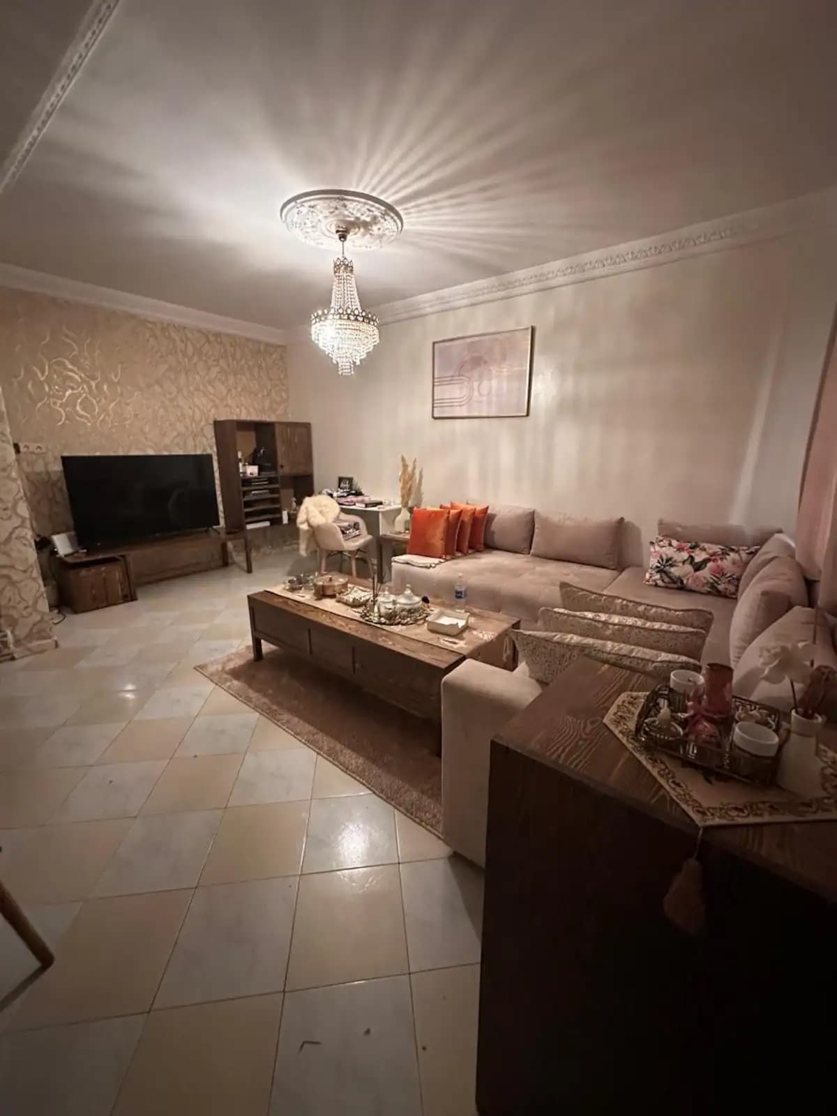 A cozy living area features a large sectional sofa adorned with patterned cushions. A decorative coffee table occupies the center, complemented by elegant accessories. Soft lighting highlights the intricate ceiling design and the warm hues of the walls, creating a serene ambiance.