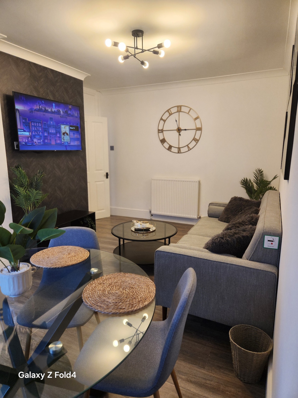 The living area features a modern light fixture above a cozy sofa, paired with decorative pillows. A television is mounted on the wall, complemented by a stylish wall clock. A glass dining table with woven placemats is seen, along with potted plants adding a touch of greenery.