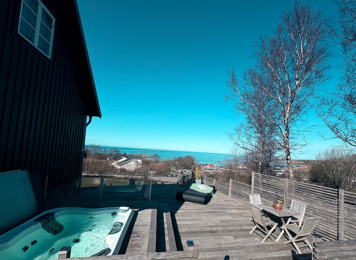 An outdoor deck features a spa pool with views of the ocean and horizon. A small dining area is set with a potted plant on the table. The surrounding area is styled with wooden fencing and trees, creating a serene environment.