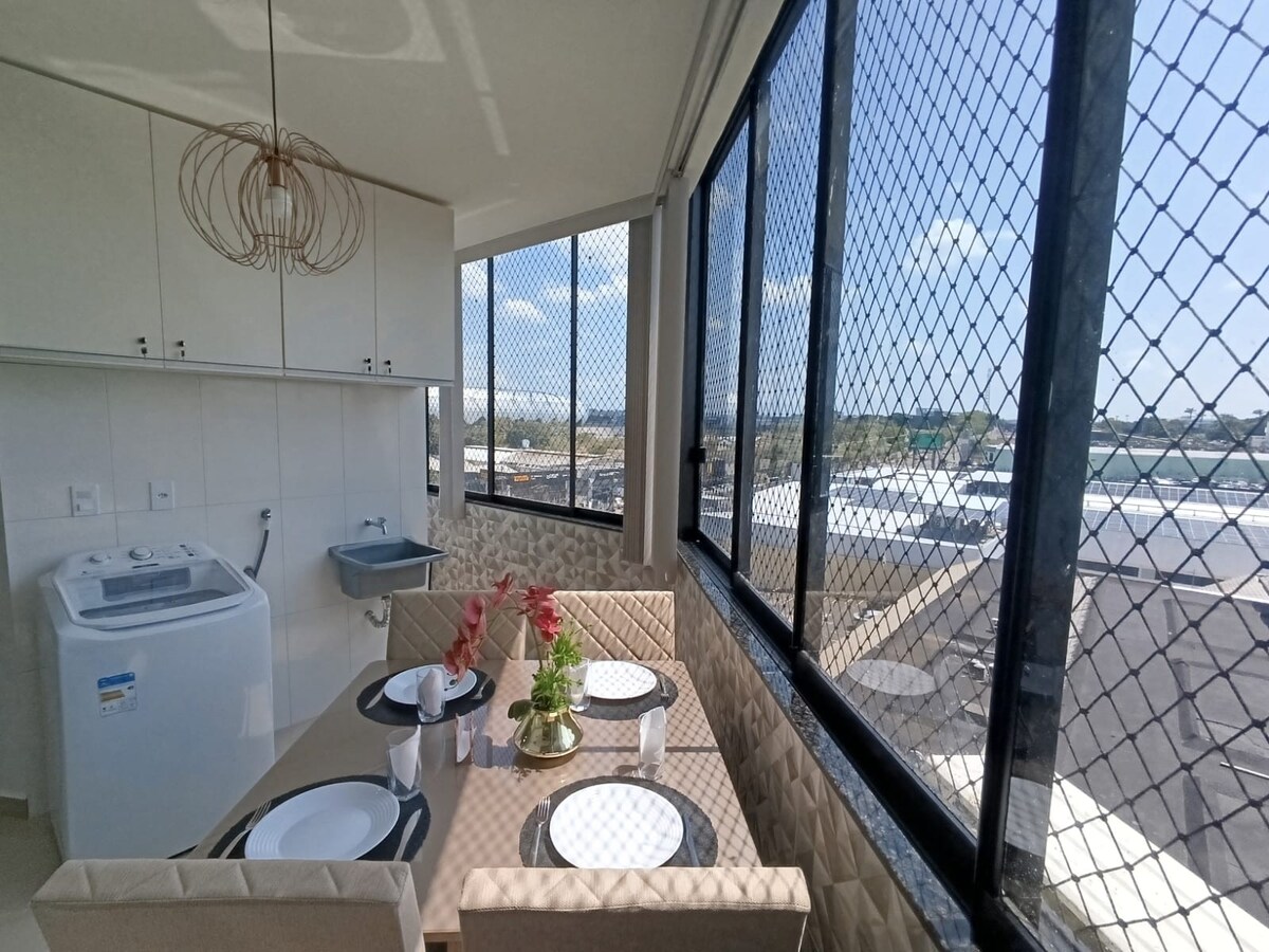 A dining area is surrounded by large windows, allowing natural light to fill the space. A round table is set for four, accompanied by a modern light fixture above. A washing machine is positioned nearby, contributing to the area’s functionality.