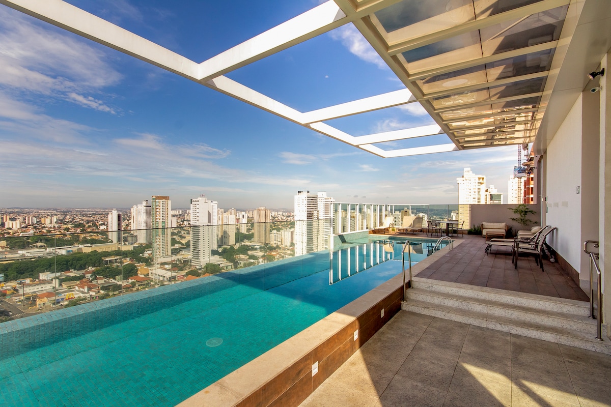 An infinity pool is viewed from a modern deck, offering panoramic city views. Lounge chairs are positioned near the pool's edge, and the structured overhang provides shaded areas. The skyline is visible against a clear blue sky, creating a serene outdoor environment.