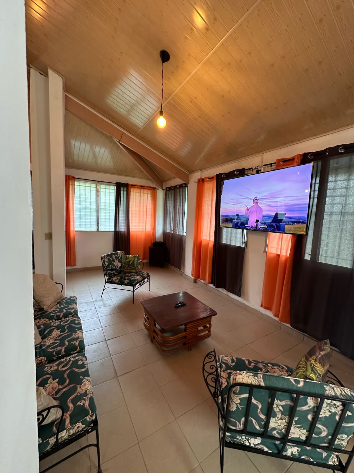 A comfortable living area features a wooden ceiling with overhead lighting. Two patterned sofas are arranged around a coffee table, and a large television is mounted on the wall. Light filters through large windows dressed with sheer and dark curtains.