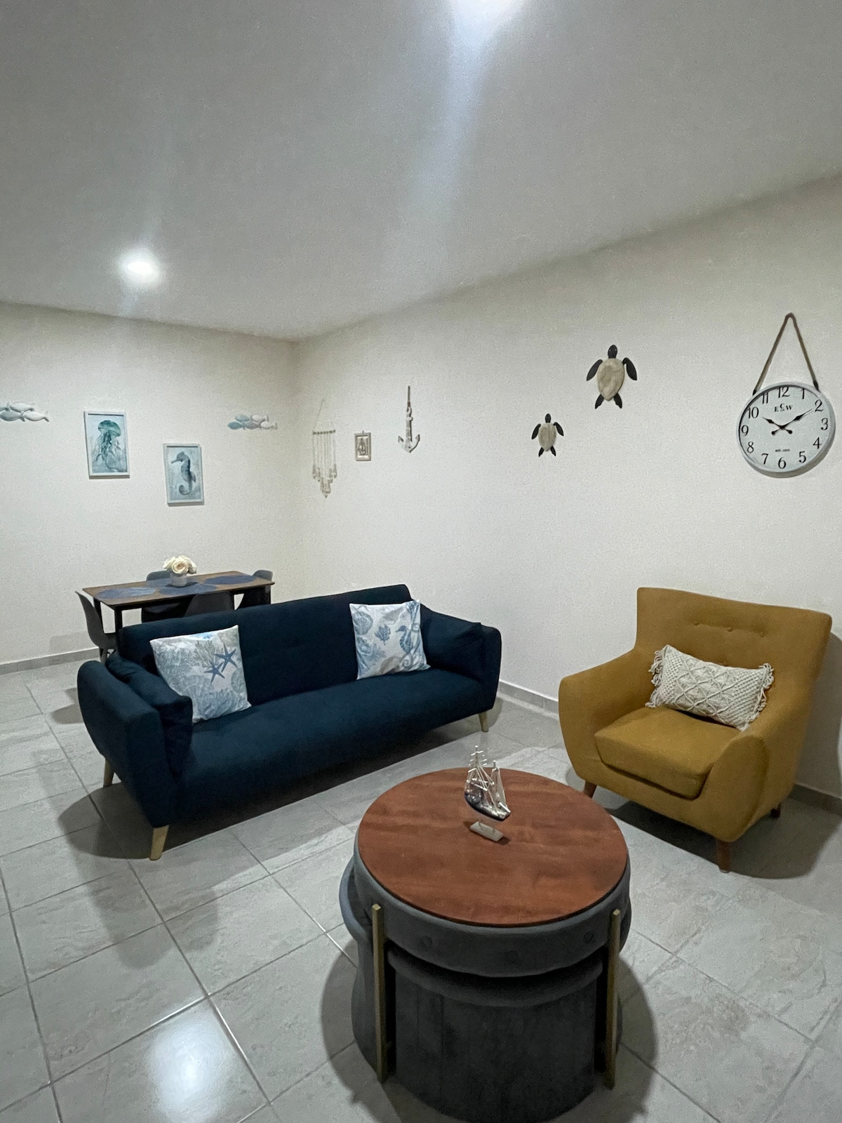 A cozy lounge area features a navy blue sofa adorned with patterned throw pillows and a mustard yellow armchair. A round wooden coffee table with decorative items occupies the center, while wall art with nautical themes decorates the light-colored walls.
