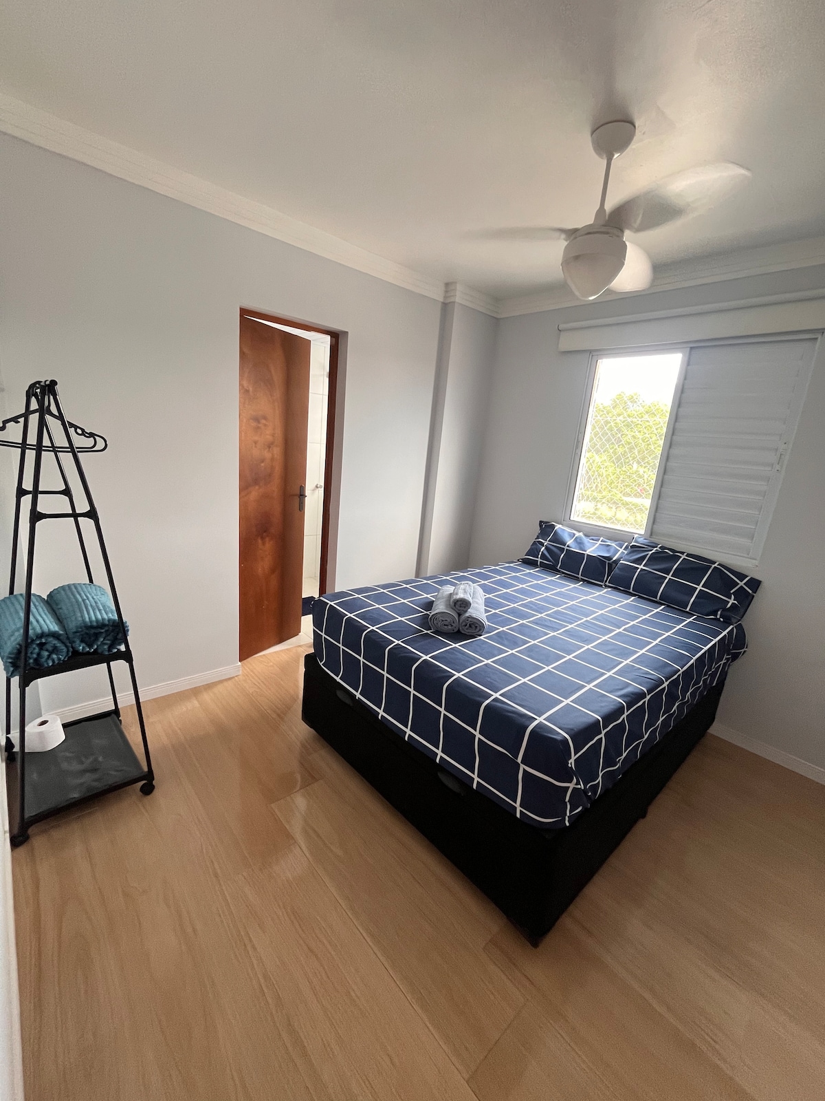 A well-lit bedroom features a spacious bed with a blue and white grid-patterned duvet. A ceiling fan hangs above, providing airflow. A window allows natural light and offers a view. A stylish clothing rack displays two rolled towels, contributing to the room's practical layout.