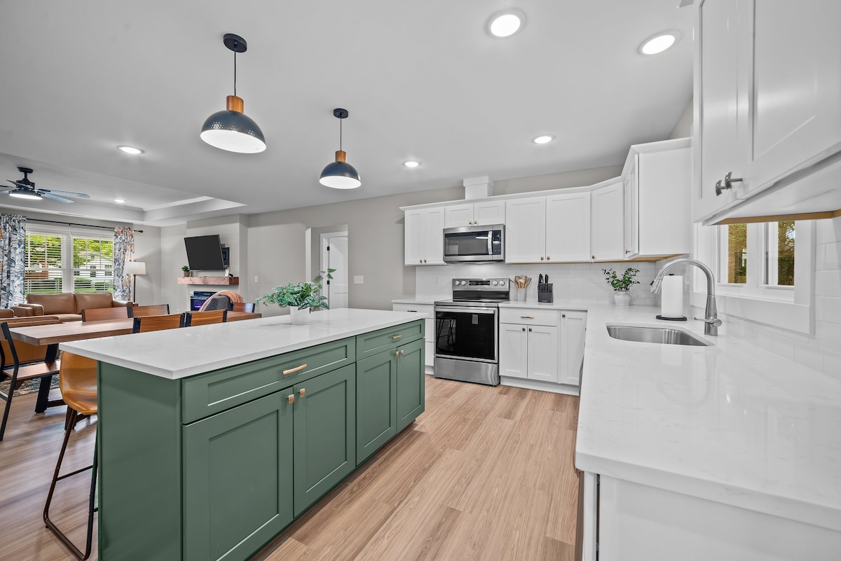 The modern kitchen features white cabinetry and dark accents, complemented by ample countertop space. New appliances, including a microwave and stove, are visible. An inviting dining area is seen in the background, illuminated by natural light from adjacent windows.