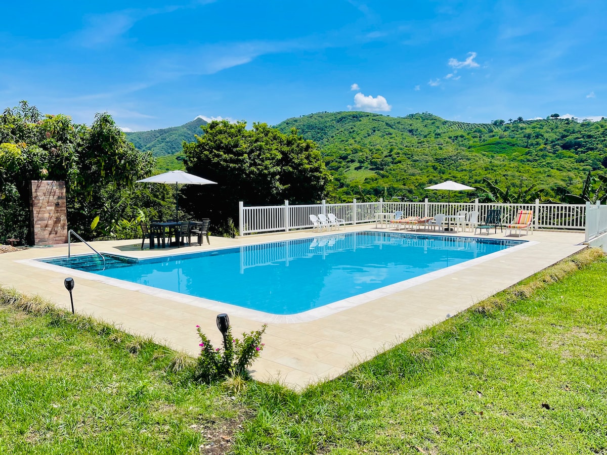 A spacious private pool is set against a backdrop of lush green mountains. Surrounded by a white fence, the pool area features sun loungers under shaded umbrellas, with a stone patio providing additional outdoor space. The clear blue water reflects the sunny sky.