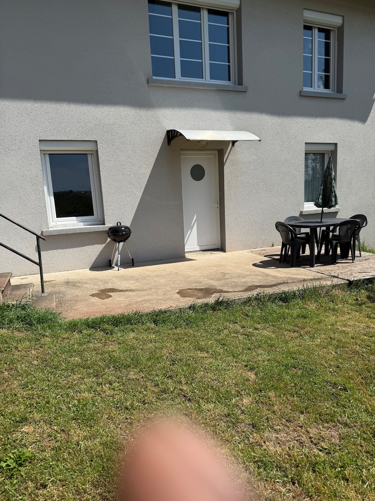 The exterior features a patio area with a small table and several black chairs, suited for outdoor dining. A door with a circular window leads inside, and a barbecue grill is positioned nearby. Lush green grass surrounds the concrete patio.