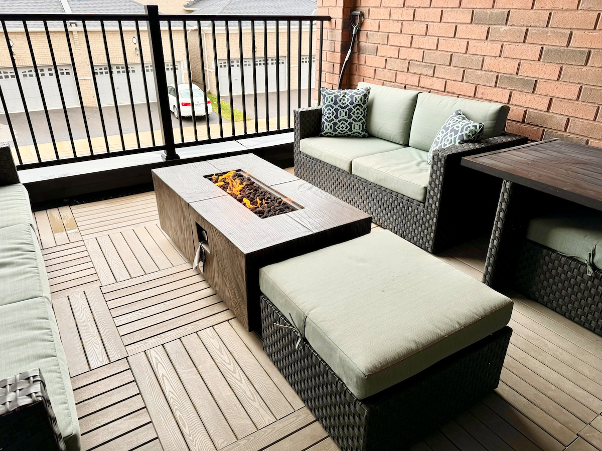 The outdoor patio features comfortable seating arrangements made of woven materials, including a sectional sofa and cushioned ottoman. A modern gas fire pit serves as the centerpiece, surrounded by stylish wooden decking and a brick wall background.