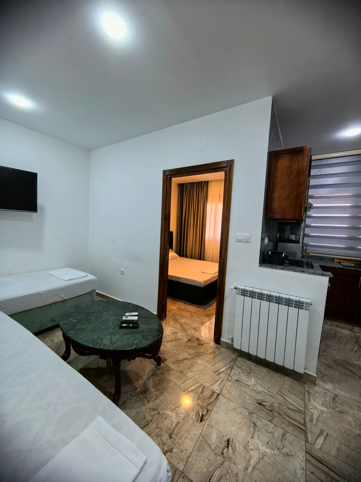 A light-filled living space is depicted, featuring two twin beds and a round table with a dark green surface. A door leads to another room, visible through the opening, while a wall-mounted television is installed in the corner. Marble flooring adds to the overall aesthetic.