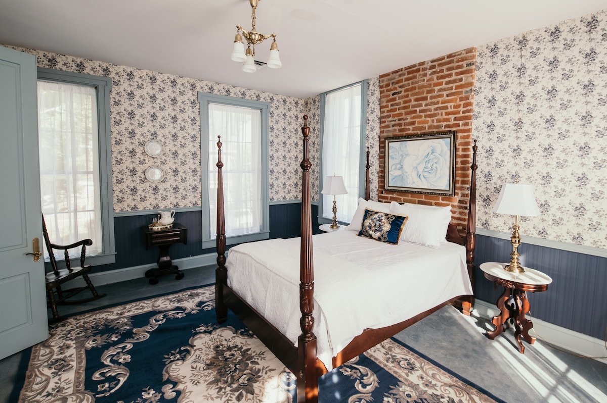 A traditional bedroom features a spacious bed adorned with white bedding. Elegant floral wallpaper complements brick accents and blue trim. A rocking chair and writing desk are positioned in the room, while two bedside tables with lamps complete the inviting atmosphere.