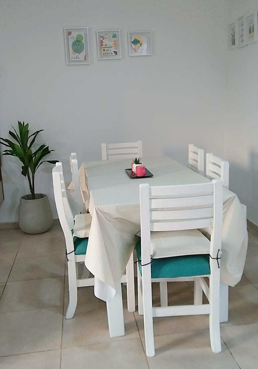 A dining area features a white table surrounded by tall white chairs with teal cushions. A small potted plant is positioned beside the table, and decorative framed art is displayed on the wall above, adding a touch of color to the neutral walls.