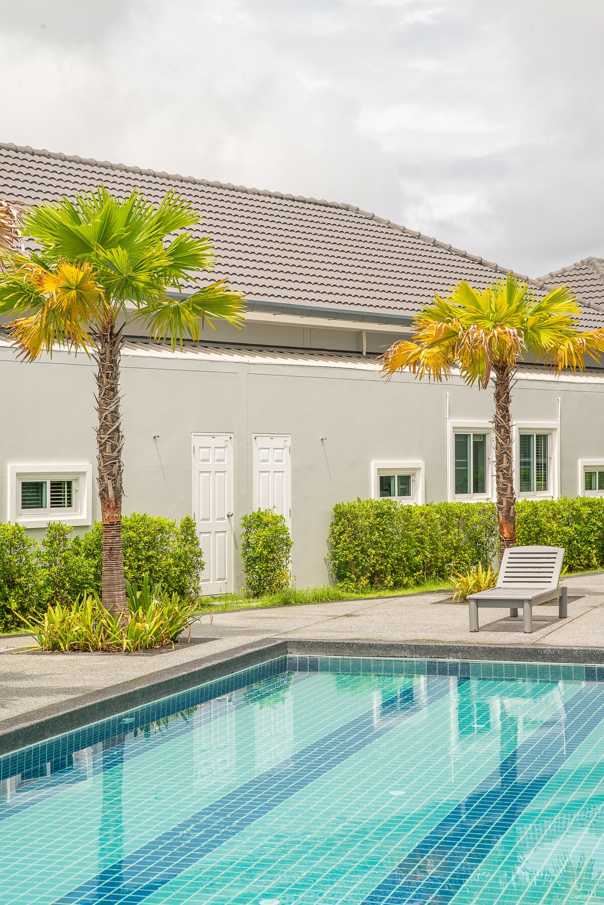 A well-maintained outdoor pool is framed by lush palm trees and neatly trimmed hedges. The building features a light-colored facade with symmetrical windows and doors, creating a serene atmosphere. A single lounge chair is positioned beside the pool, inviting relaxation.