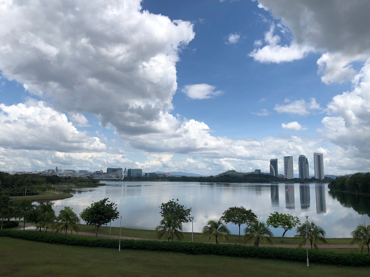 A scenic view of Putrajaya Lake reflects the sky, with lush greenery lining the shore. The background features modern buildings in varying heights against a backdrop of clouds and blue sky, creating a tranquil lakeside atmosphere.